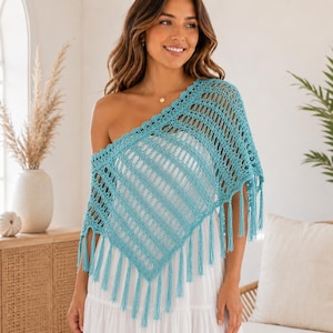 Könnte beinhalten: Ein türkisfarbener Häkelponcho mit durchbrochenem Rautenmuster und langen Fransen an den unteren Kanten. Das Kleidungsstück wird über einem weißen Kleid getragen und zeigt einen diagonalen Fall, der eine Schulter mit gewelltem Ausschnitt freilässt.