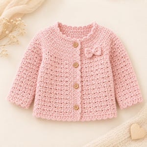 May include: A light pink long-sleeve cardigan made of crocheted yarn. The garment features a scalloped edge along the neckline, cuffs, and front opening. It is fastened with four round wooden buttons and includes a small crocheted bow detail.