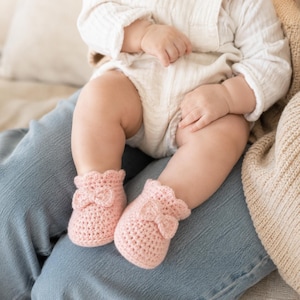 May include: A pair of soft, light pink crocheted baby booties featuring a textured knit pattern, a small decorative bow on the front of each bootie, and a scalloped edge around the ankle opening, designed for infants with a cozy, handcrafted look.