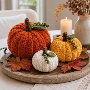 May include: Three crocheted pumpkins in orange, yellow, and white, each featuring a brown stem and green leaves. The pumpkins are arranged on a round wooden tray alongside scattered dried maple leaves, creating a textured autumn display.