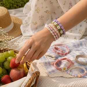 May include: A collection of handmade crochet bracelets featuring floral patterns, daisy motifs, and pastel-colored beads. The bracelets are crafted with intricate thread work in shades of purple, white, pink, and green, displayed alongside fresh fruit.