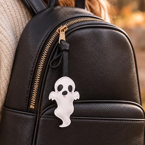 Könnte beinhalten: Ein schwarzer Mini-Rucksack aus Kunstleder mit goldenem Reißverschluss und Tragegriff. Ein weißer, geisterförmiger Anhänger mit schwarzen Augen und kleinen rosa Wangen ist am vorderen Reißverschluss befestigt und hat genähte Kanten.