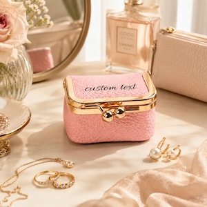Puede incluir: Un pequeño joyero rectangular de color rosa con textura, marco de metal dorado y cierre de broche. La parte frontal de la caja presenta las palabras custom text en una fuente cursiva negra. La caja tiene una forma estructurada y superficie granulada.