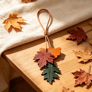 Puede incluir: Un adorno colgante decorativo que presenta tres recortes distintos en forma de hoja hechos de cuero en tonos verde oscuro, marrón rojizo y naranja. Las hojas están unidas a un cordón de cuero fino y en bucle que está anudado en la parte superior.