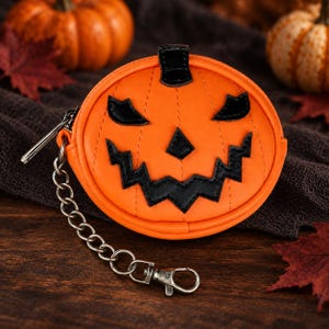 May include: A round orange coin purse shaped like a jack-o'-lantern with a black stitched face featuring triangular eyes, a triangular nose, and a jagged mouth. It includes a black stem at the top and a silver metal chain with a lobster claw clasp.