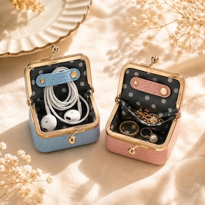 May include: Two small square cases with gold-tone metal frames and kiss-lock closures. One case is light blue, the other is pink. Both feature a grey interior with white polka dots, a snap-button strap, and a small pocket for storing items.