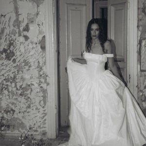 May include: A floor-length white gown featuring an off-the-shoulder neckline with gathered fabric details, a fitted bodice, and a voluminous, pleated skirt that extends into a train, captured in a black and white photograph.