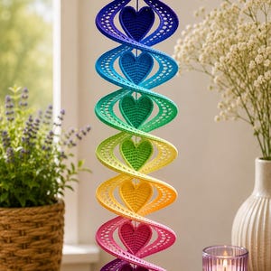 Peut inclure: Un mobile décoratif en spirale suspendu composé d'une séquence verticale de six cœurs aux couleurs de l'arc-en-ciel : violet, bleu, cyan, vert, jaune, orange et rose. La structure présente une texture perforée avec un motif en treillis.