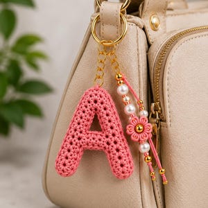 Puede incluir: Un llavero con la letra A tejida a ganchillo en color rosa, sujeto a una anilla de metal dorado. El llavero incluye un colgante con perlas blancas, pequeñas cuentas doradas y una flor hecha de cordón rosa que cuelga de una cadena dorada.