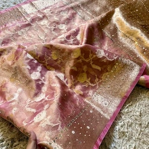 May include: A pink and gold sari featuring an intricate floral pattern woven into the fabric. The garment includes a wide, metallic gold border embellished with small white beads and reflective mirror accents throughout the decorative trim.
