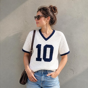 May include: A short-sleeved, V-neck crochet top in off-white with dark navy blue trim on the neckline and sleeve cuffs. The front of the garment features the number 10 in large, dark navy blue block lettering centred on the chest area.