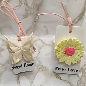 Può includere: Due ornamenti rettangolari bianchi da appendere con cordini rosa. L'ornamento a sinistra presenta una farfalla color crema e il testo "Sweet Home" in nero. L'ornamento a destra mostra una margherita gialla con centro rosa e il testo "True Love".