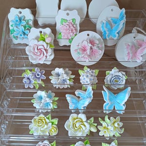 Può includere: Una collezione di ornamenti in ceramica bianca con motivi floreali e farfalle in rilievo nei toni del rosa, blu, giallo e verde. Gli ornamenti sono esposti su un supporto a gradini in acrilico trasparente, con varie forme tra cui cerchi e ovali.