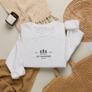 Könnte beinhalten: Ein hellgraues Sweatshirt mit Rundhalsausschnitt und einer schwarzen Grafik von drei Tannenbäumen, darunter der Text ESTD 1989, THE WANDERER und Melanie in einer klaren, serifenlosen Schrift mittig auf der Brust.