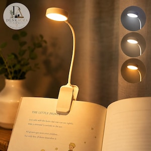 May include: A white clip-on reading lamp with a flexible neck and a circular light head, attached to an open book. The lamp features three adjustable color temperature settings, ranging from cool white to warm yellow light, as shown in the circular insets.
