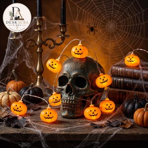 Op de afbeelding: Een lichtsnoer met oranje LED-pompoenen in de vorm van jack-o'-lanterns, gedrapeerd over een donkere, verweerde menselijke schedel. De kleine, gloeiende pompoenlampjes hebben zwarte uitgesneden gezichten en zijn verbonden door een dunne draad.