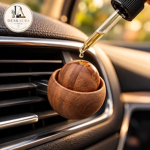 Puede incluir: Un difusor de ambientador esférico de madera para coche fijado a una rejilla de ventilación. Un cuentagotas de cristal con tapón negro se sitúa sobre la esfera de madera, liberando una gota de líquido dorado sobre la superficie de madera marrón.