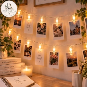 May include: A string of warm white LED fairy lights featuring clear plastic clips to hold photographs. The lights are arranged in two horizontal rows, illuminating various printed images including landscapes, flowers, and text-based quotes.