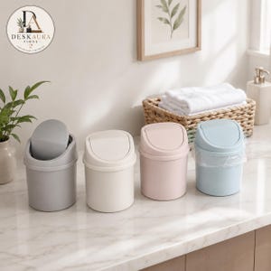 Puede incluir: Cuatro cubos de basura cilíndricos pequeños de plástico con tapas basculantes, dispuestos en fila. Los colores son gris, blanco roto, rosa claro y azul claro. El cubo azul claro contiene una bolsa de plástico transparente.