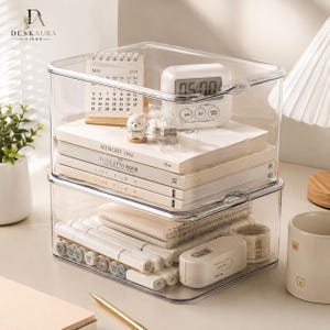 May include: A two-tier clear acrylic stackable storage organiser with drawers. The top drawer holds a May 2024 calendar, a white digital clock, and books titled Memory 1981, The Stiletto Book, and Life Film. The bottom drawer contains markers and tape.