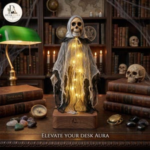 May include: A skull-headed figure draped in layered, tattered grey and black fabric, illuminated by internal warm string lights. The figure stands on a rectangular wooden base with a logo, surrounded by vintage books, a compass, and stones.