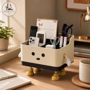 Op de afbeelding: Een crèmekleurige rechthoekige bureau-organizer met een zwarte basis, vier kleine gele voetjes en twee witte armen. De voorkant heeft een eenvoudig gezicht met twee zwarte ogen en een kleine zwarte mond. Het bevat een smartphone, afstandsbediening, kalender en pennen.