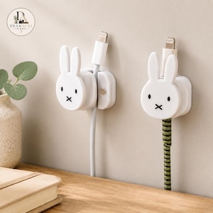 Puede incluir: Dos organizadores de cables de pared blancos con forma de cabeza de conejo, con ojos negros y una boca en forma de cruz. Cada organizador sujeta un cable de carga; uno es blanco liso y el otro tiene un patrón de espiral verde y negro.