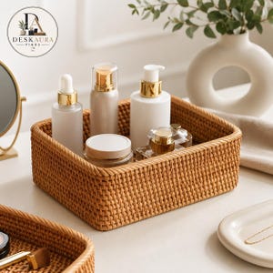May include: A rectangular woven rattan storage basket containing several skincare bottles and jars with gold-toned pumps and lids. The basket features a tight, uniform weave pattern and sits alongside a white ceramic vase and a small tray.