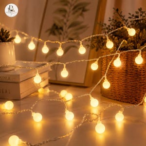 May include: A string of decorative globe-shaped LED lights emitting a warm, soft glow. The small, spherical bulbs are evenly spaced along a thin, flexible wire, creating a luminous effect that illuminates the surrounding space with golden light.