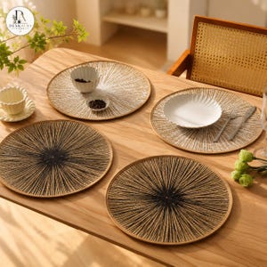 May include: Four round placemats featuring a sunburst pattern of radiating lines. Two placemats have a white center fading to tan, and two have a black center fading to tan. The placemats are arranged on a light wood table with white ceramic dishes.