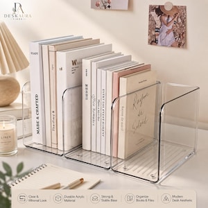 May include: A clear acrylic book organizer featuring three connected U-shaped compartments. The organizer holds several books with various white and beige spines, including titles like Made & Crafted, Office Romance, and Rayon de Soleil, on a flat surface.