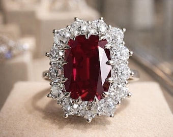 Puede incluir: Un anillo que presenta una gran piedra preciosa rectangular de color rojo intenso en el centro, rodeada por un halo de doce piedras blancas redondas, claras y facetadas engastadas en una banda metálica. El diseño resalta el contraste entre la piedra roja central y las piedras blancas circundantes.