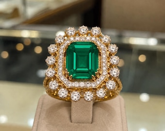 Puede incluir: Un anillo de oro con una gran piedra preciosa verde de corte esmeralda rectangular en el centro. La piedra está rodeada por un doble halo de pequeños diamantes blancos redondos y claros, con un borde exterior festoneado con diamantes.