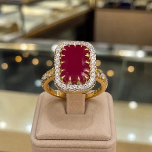 Puede incluir: Un anillo de oro con una gran piedra preciosa rectangular de color rojo intenso engastada con garras. La piedra central está rodeada por un halo de pequeños diamantes redondos transparentes. El aro de oro pulido también tiene diamantes laterales.