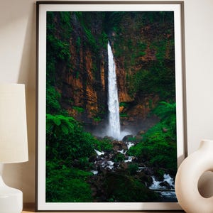 May include: A framed wall art print featuring a tall, narrow waterfall cascading down a dark, mossy cliff face surrounded by lush green vegetation and rocky terrain, presented in a thin black frame on a wooden surface with a lamp and vase.
