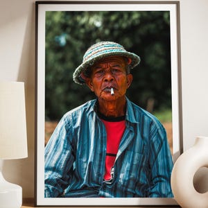 May include: A framed wall art print featuring a portrait of an older person wearing a textured, multi-coloured bucket hat and a blue striped button-down shirt over a red t-shirt, with a cigarette held between their lips against a dark background.