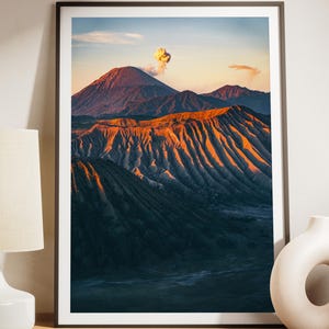 Puede incluir: Una impresión de paisaje enmarcada que muestra una cordillera volcánica al amanecer con luz dorada en las laderas estriadas y una pequeña columna de humo en la cima. El texto NO IA ORIGINAL PHOTO MADE BY HUMAINS DIGITAL DOWNLOAD es visible.