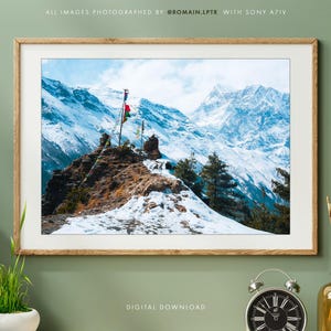 Pode incluir: Uma fotografia de paisagem emoldurada apresentando uma cordilheira coberta de neve com um primeiro plano rochoso e bandeiras de oração coloridas. A impressão está em uma moldura de madeira clara, com uma pequena planta e um despertador.