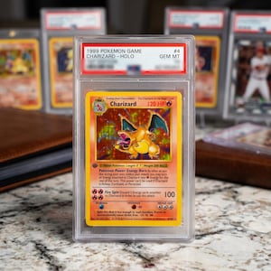 May include: A 1999 Pokemon Game Charizard holographic trading card encased in a clear protective slab. The card features a yellow border, a fire-type Charizard illustration, 120 HP, and the text 1999 POKEMON GAME CHARIZARD - HOLO GEM MT #4.