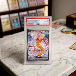 Puede incluir: Una tarjeta coleccionable Pokemon M2a JP 2025 #246 graduada que presenta una colorida ilustración de una criatura naranja similar a un dragón. La tarjeta está encerrada en una funda protectora de plástico transparente y rectangular.