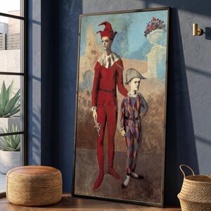 May include: A framed vertical wall art print depicting a tall figure in a solid red jester costume with a matching cap and a shorter figure in a blue and red diamond-patterned harlequin suit with a grey hat, set against a muted outdoor landscape.