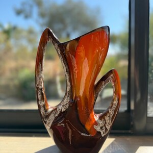 Może przedstawiać: Ceramiczny wazon z błyszczącą, ciemnobrązową podstawą, która przechodzi w żywą, pomarańczowo-żółtą szkliwioną górną część. Wazon posiada dwa duże, ażurowe uchwyty z teksturowanym, białym, gąbczastym wykończeniem dekoracyjnym wzdłuż krawędzi.