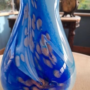 Può includere: Un vaso in vetro caratterizzato da un motivo a vortice nei colori blu scuro, azzurro e bianco. La superficie presenta una finitura liscia e lucida con pagliuzze metalliche dorate incorporate che creano un effetto maculato nel corpo in vetro.