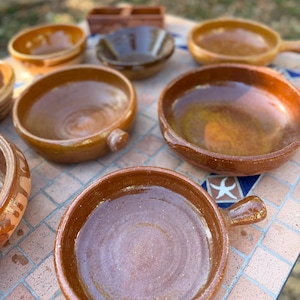 Pode incluir: Uma coleção de pratos e taças de terracota vidrada, redondos e em vários tons de castanho. O conjunto inclui várias frigideiras rasas com pegas pequenas, taças fundas e um recipiente retangular dividido, todos com um acabamento liso.