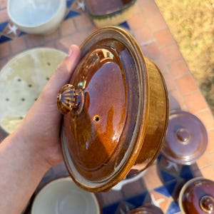 Peut inclure: Un plat à four rond en céramique émaillée marron avec un couvercle assorti doté d'un petit bouton de préhension et d'un évent à vapeur. La finition brillante reflète la lumière, soulignant la surface lisse et incurvée de cet ustensile.
