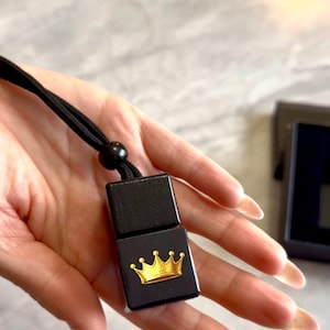 May include: A black square-shaped pendant featuring a gold crown graphic on the front. The pendant is attached to a black cord with a small black bead accent. The design is simple and geometric, showcasing a smooth finish on the black surface.