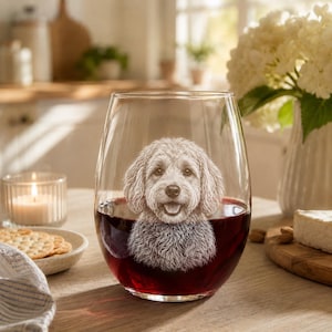 May include: A clear stemless wine glass filled with red wine, featuring a detailed white etched illustration of a curly-haired dog on the front surface. The dog has floppy ears, a wide smile, and a textured coat, centered on the glass.