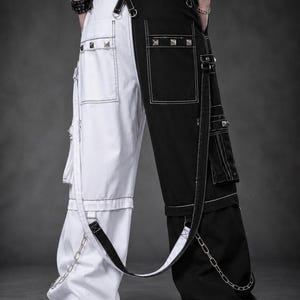 May include: Split-leg cargo pants featuring a white left leg and a black right leg. The pants include multiple pockets with silver-toned pyramid studs, white contrast stitching, adjustable straps, and silver metal chains attached to the lower legs.