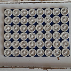 May include: A collection of forty-eight circular buttons arranged in a grid on a dark blue rectangular backing. Each button features a white thread-wrapped design with a central point radiating outward to the circular edge of the button frame.