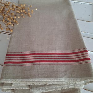 Könnte beinhalten: Gefaltetes beiges Küchenhandtuch aus Leinen mit drei roten Streifen. Das Handtuch besteht aus einem Naturstoff und hat einen rustikalen, ländlichen Stil. Das Handtuch ist gefaltet und gestapelt.